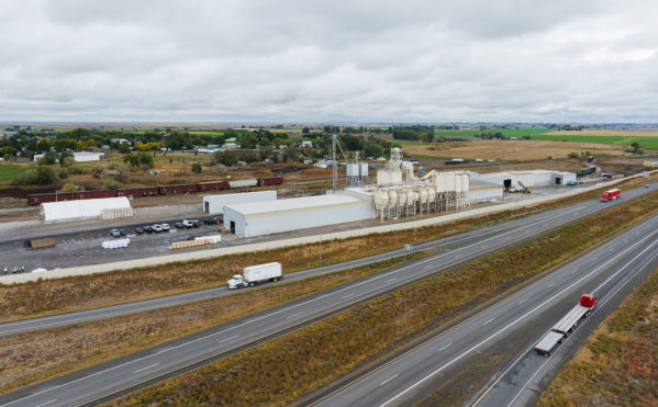 Aerial of JDH mill in Wendell, Idaho. The mill is next to a highway with semis driving. You can see a vast amount of farmland in the background. the sky is gray and cloudy.